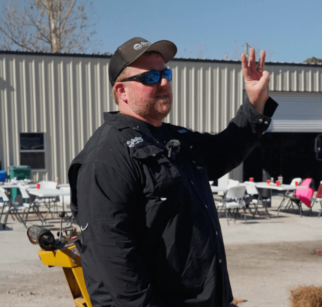 A person wearing sunglasses and a cap gestures outside near industrial buildings and tables set up with chairs, in a sunny setting.