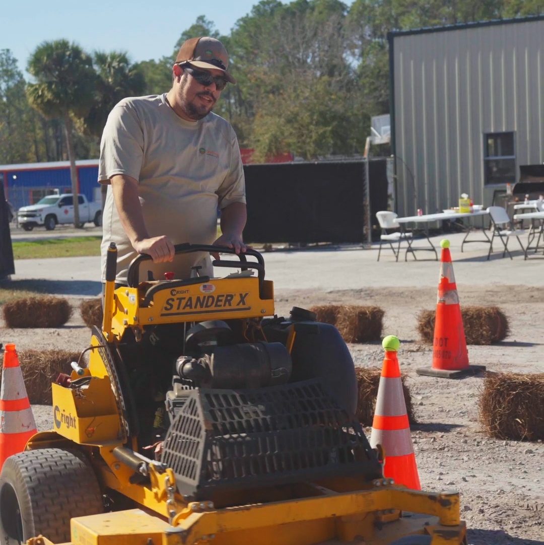 A person operates a yellow Stander X mower in a parking lot with traffic cones and hay bales, surrounded by outdoor tables and trees.