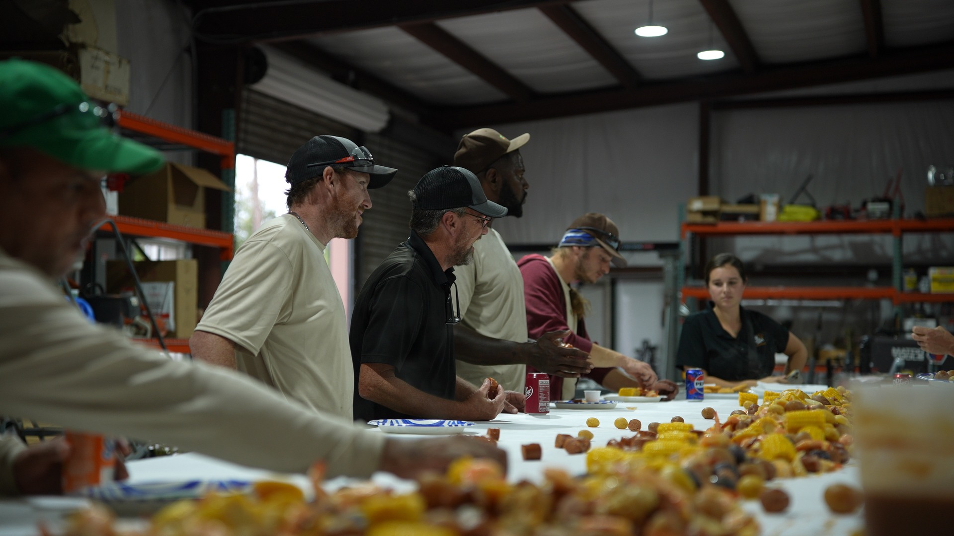 A group of people gather around a table covered with corn and sausages inside a warehouse. Industrial shelves line the walls.