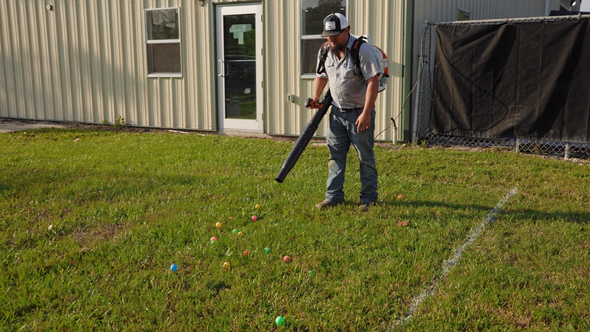 A person using a leaf blower stands on grass beside a building, surrounded by scattered multicolored balls.